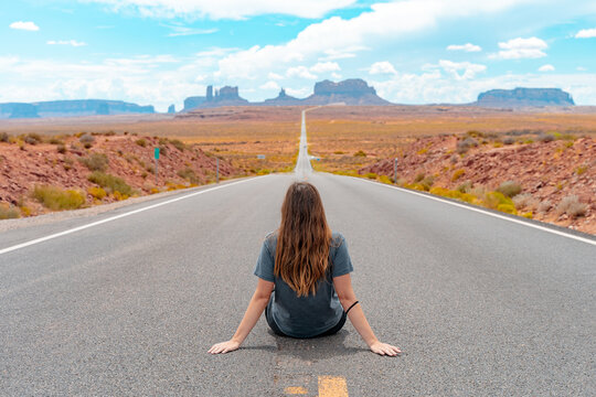 Attractive young woman from back view sitting in the middle of American straight road in the desert.