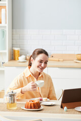 Fototapeta na wymiar Vertical portrait of smiling woman enjoying breakfast in morning and using digital tablet in kitchen