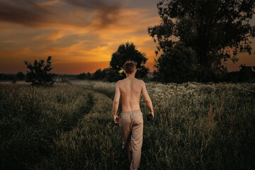 man with a naked torso walks along a half-way road in the grass at sunset, the concept of youth and travel, loneliness.