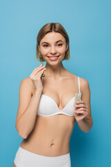 Fototapeta premium cheerful young woman in white bra holding jade scrapper and roller isolated on blue