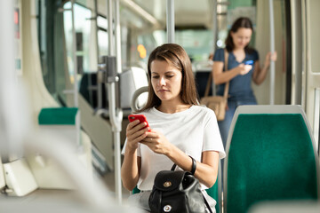 Fototapeta premium Young woman browsing and typing messages on phone on way to work in modern streetcar ..