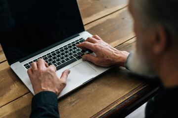 Close-up selective focus of unrecognizable bearded mature older male typing on laptop keyboard sitting at table at home office. Closeup of aged business man working on computer indoors, surfing online