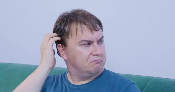 Portrait of young man with dissatisfied look, examining something new and unusual and is grimacing at this, being distrustful of changes. Then he thinks about it and decides to try, scratching head