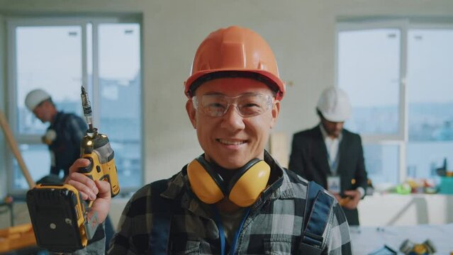 Close up builders asian man wearing protective helmet and goggles look at camera smiling holds screwdriver. On background foreman and architect discussing plan. Portrait. Slow motion