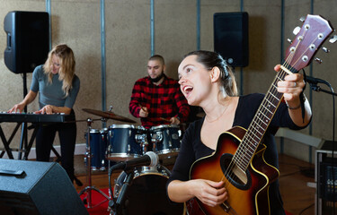 Attractive smiling female soloist playing guitar and singing with her music band in sound studio