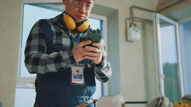 Close up face young builder man in hardhat and goggles working using smartphone. On table blueprints during renovation in building. Slow motion