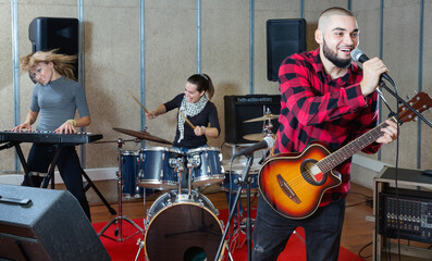 Rehearsal of music band. Positive smiling guy guitar player and singer practicing with band members in recording studio.