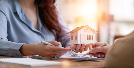 Cropped image of real estate agent assisting client to sign contract at desk with house model.