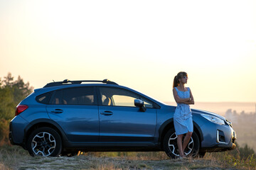 Happy young woman driver in blue dress enjoying warm summer evening standing beside her car. Travelling and vacation concept