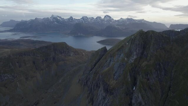 Aerial view of steep mountains with snowy peaks in the background, in Lofoten, Norway - reverse, tilt, drone shot