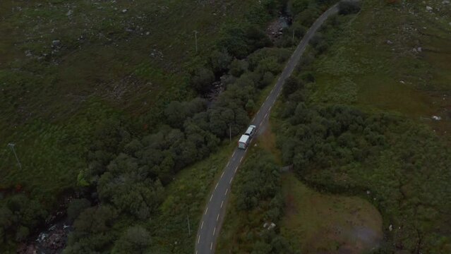 Forwards tracking of car with horse trailer driving on country road along stream. High angle view of valley with trees and shrubs. Ireland