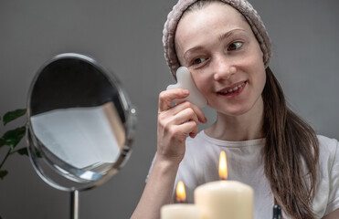 Woman is looking in the mirror and doing a massage with a gouache scraper. Candles and an aroma lamp with essential oils on the table. Concept of facial care, aromatherapy, morning and evening rituals