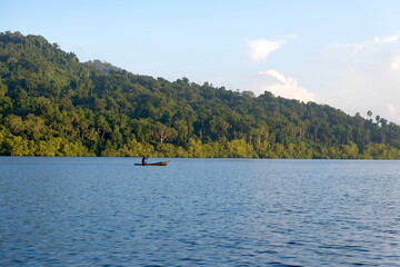 A person paddling a traditional fishing dugout canoe in the ocean with tree covered tropical island...