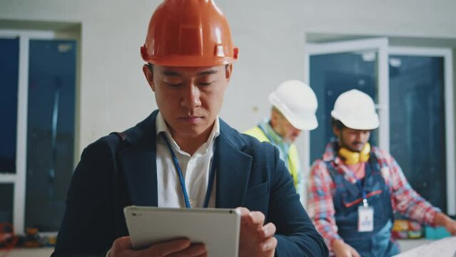Young asian engineer, architect wearing a white protective helmet in a suit use tablet computer stand look around. On bakground builders in hardhats working in building. Slow motion