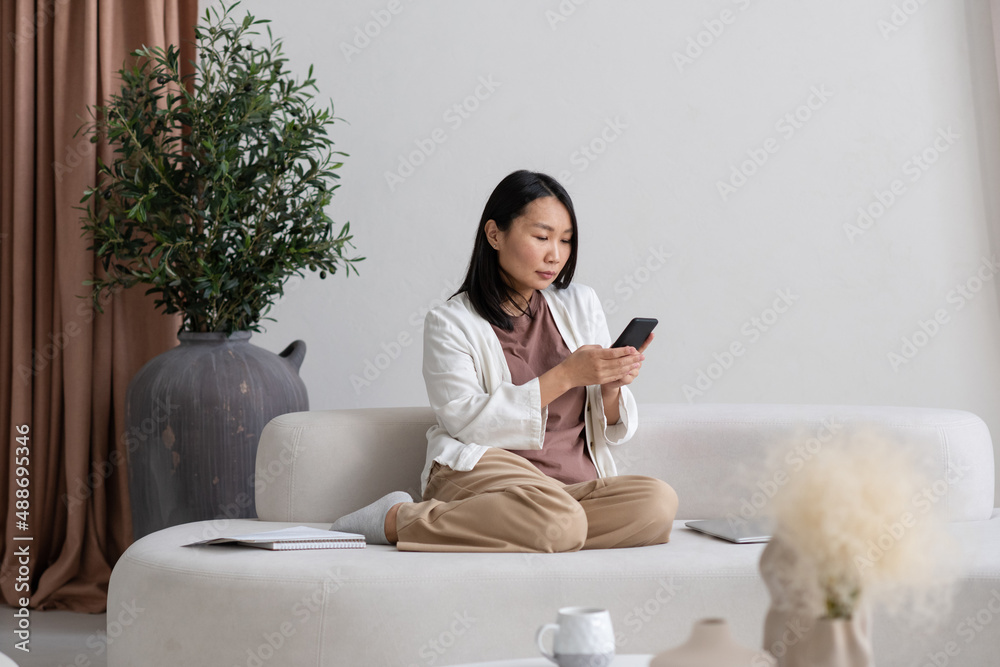 Aufkleber Young serious freelancer with smartphone texting or searching in the net while sitting on sofa against large vase with green domestic plant