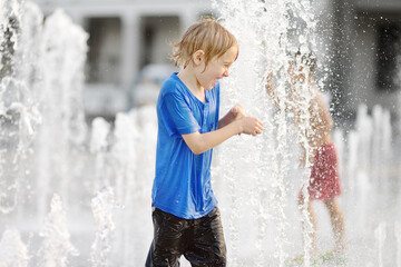 Little boy plays in the square between the water jets in the dry fountain at sunny summer day. Active leisure for kids in the city. - obrazy, fototapety, plakaty