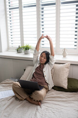 Obraz premium Young tired employee with laptop on crossed legs stretching raised arms while sitting on double bed and working online from home office