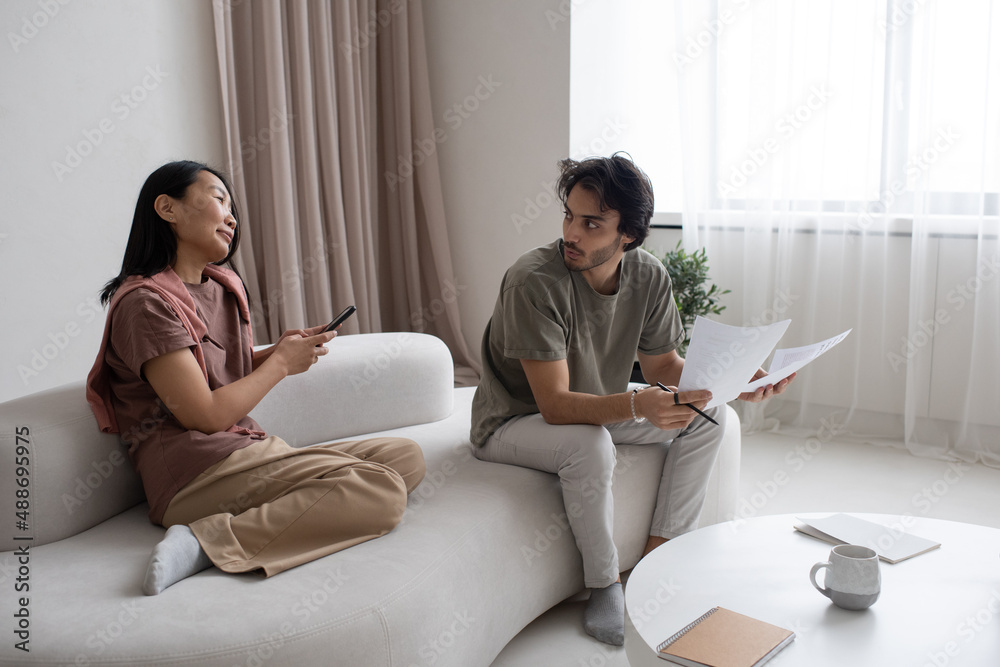 Poster Two young intercultural employees having discussion of working points while analyzing papers with financial data in home office