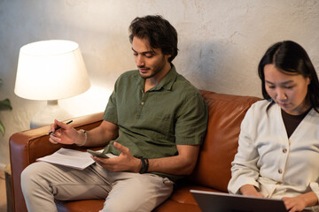 Two young intercultural employees in casualwear using wireless gadgets while sitting on leather couch and analyzing financial data