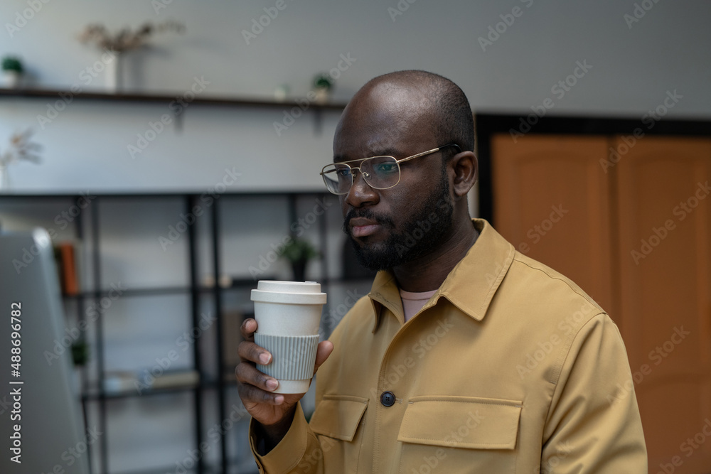 Poster Young serious office manager with drink looking at data on computer screen while sitting in front of monitor by workplace