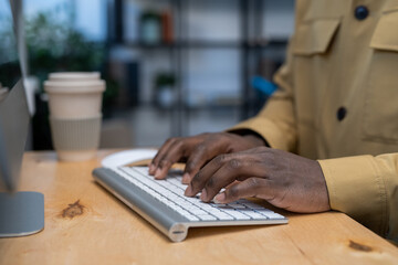 Hands of young African-American businessman pressing keys of computer keyboard while working in the internet by wooden table