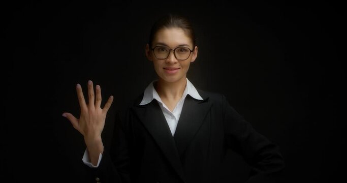 Asian woman in glasses and business clothes, smiling, shows five fingers with her right hand. Businesswoman counts with her fingers on her hand. Isolated on a black background.