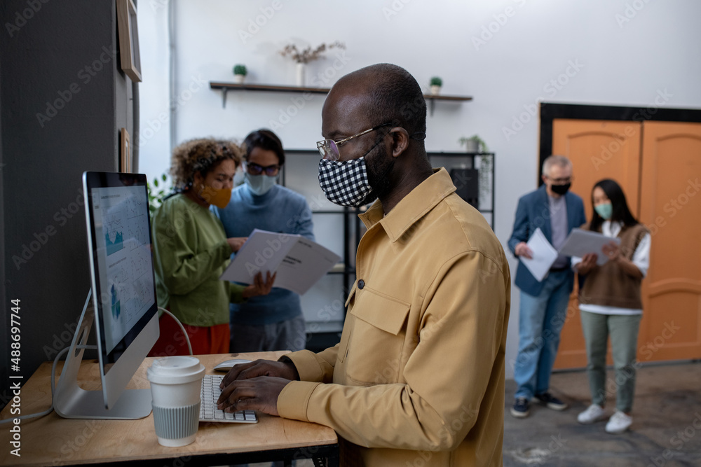 Aufkleber Young African-American economist analyzing data on computer screen while standing by desk in front of monitor with colleagues on background