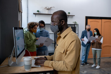Young African-American economist analyzing data on computer screen while standing by desk in front of monitor with colleagues on background
