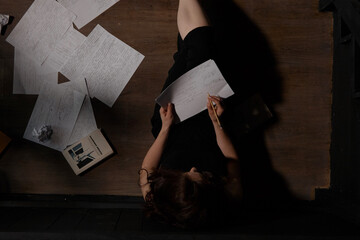 A woman in black clothes is sitting on the floor, sheets are scattered next to her. The process of writing a text, a creative crisis.