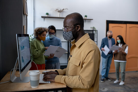 Young African-American economist analyzing data on computer screen while standing by desk in front of monitor with colleagues on background