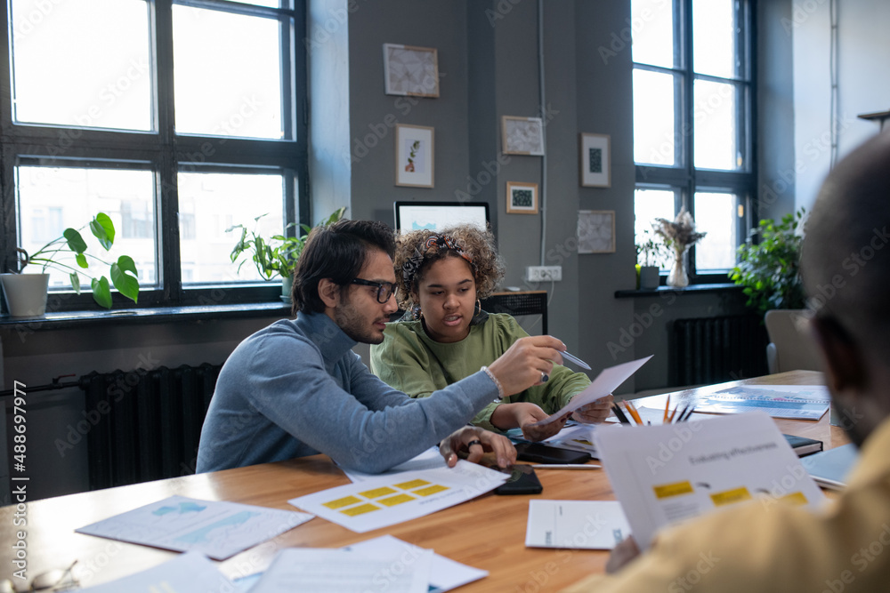 Poster Young man pointing at paper while explaining data to biracial colleague at working meeting while sitting by table in front of coworker