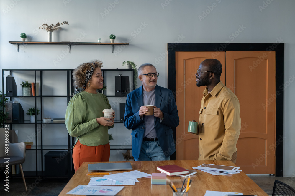 Poster three confident intercultural colleagues having drinks and talking at break while standing by large 