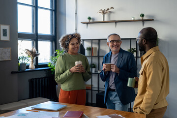 Three successful colleagues in smart casualwear having drinks at break by workplace in office while...