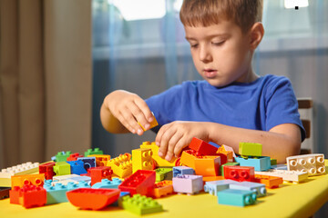 Fototapeta premium A beautiful boy is playing at home with building blocks. A cute smiling boy is playing with a constructor with a lot of colorful plastic blocks in the room, building a city. Preschool classes.