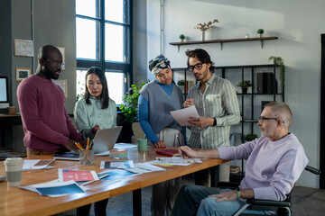 Large group of intercultural colleagues discussing online financial data and papers while standing by workplace in urban office