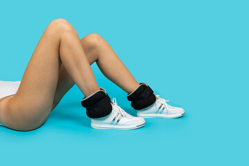 Closeup of woman wearing white bodysuit and sneakers with ankle weights lying on blue background doing physical exercises. Healthy lifestyle, working out. 