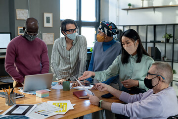 Contemporary intercultural employees in protective masks discussing financial data at meeting while gathered around table