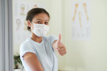Girl in mask gesturing with thumb up and showing that she ok after vaccination while standing at hospital