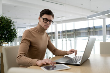 Young serious high-tech developer using tablet and laptop while sitting by tabel and working over new programming project