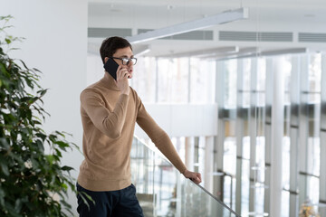 Young confident ceo in smart casualwear standing in office building and holding mobile phone by ear while consulting client of company