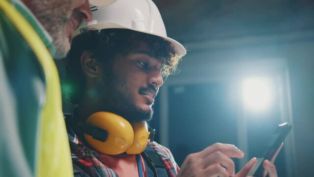 Attractive foreman and builder men in hardhats working talking using smartphone in the evening light. On table blueprints during renovation in building. Slow motion
