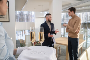 Young waitress in white gloves carrying tray with two flutes of champagne for employees discussing working points before business event