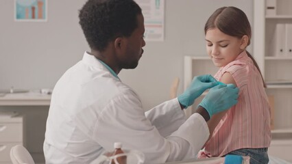 Medium slowmo shot of young African-American doctor putting band aid on shoulder of Caucasian schoolgirl after doing Covid-19 vaccine injection