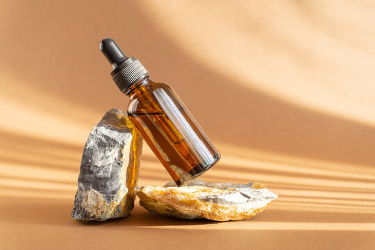 Serum in glass amber bottle with dropper lid on stand made of natural granite stone. Essential oil for care of women's skin on brown background, in rays of sunlight. Concept of beauty, rejuvenation