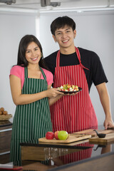 Portrait shot of Asian young lover couple girlfriend and boyfriend wear apron standing smiling look at camera helping each other slice fruits on wooden cutting board on kitchen counter table at home
