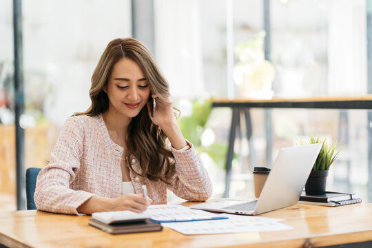 Young happy attractive beautiful asia female staff laugh talk on mobile phone relax comfort sit work on desk table at home. Remotely work on distance job telesale or secretary career for business.