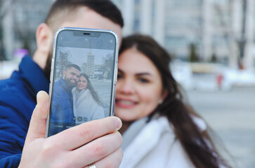 Love story of couple walking in city. Young business man in a blue jacket with a beard. And a cute woman in long coat. talking on the phone, making selfie. Bloggers. Family holiday, date