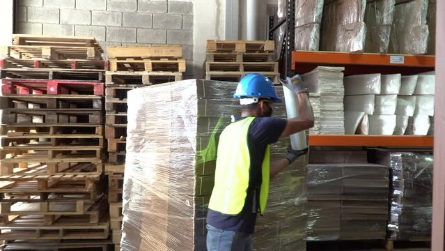 A male worker inside a warehouse, covering a pallet with a roll of transparent plastic wrap. Handheld pedestal shot.
