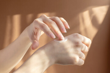 Application of cream, lotion on hands. Beautiful female hands rub moisturizing liquid into delicate skin, in rays of sunlight on brown background. Concept of care and beauty
