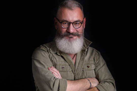 Mature bearded caucasian man looks slyly in camera with eyeglasses on his face, military style green shirt, black background